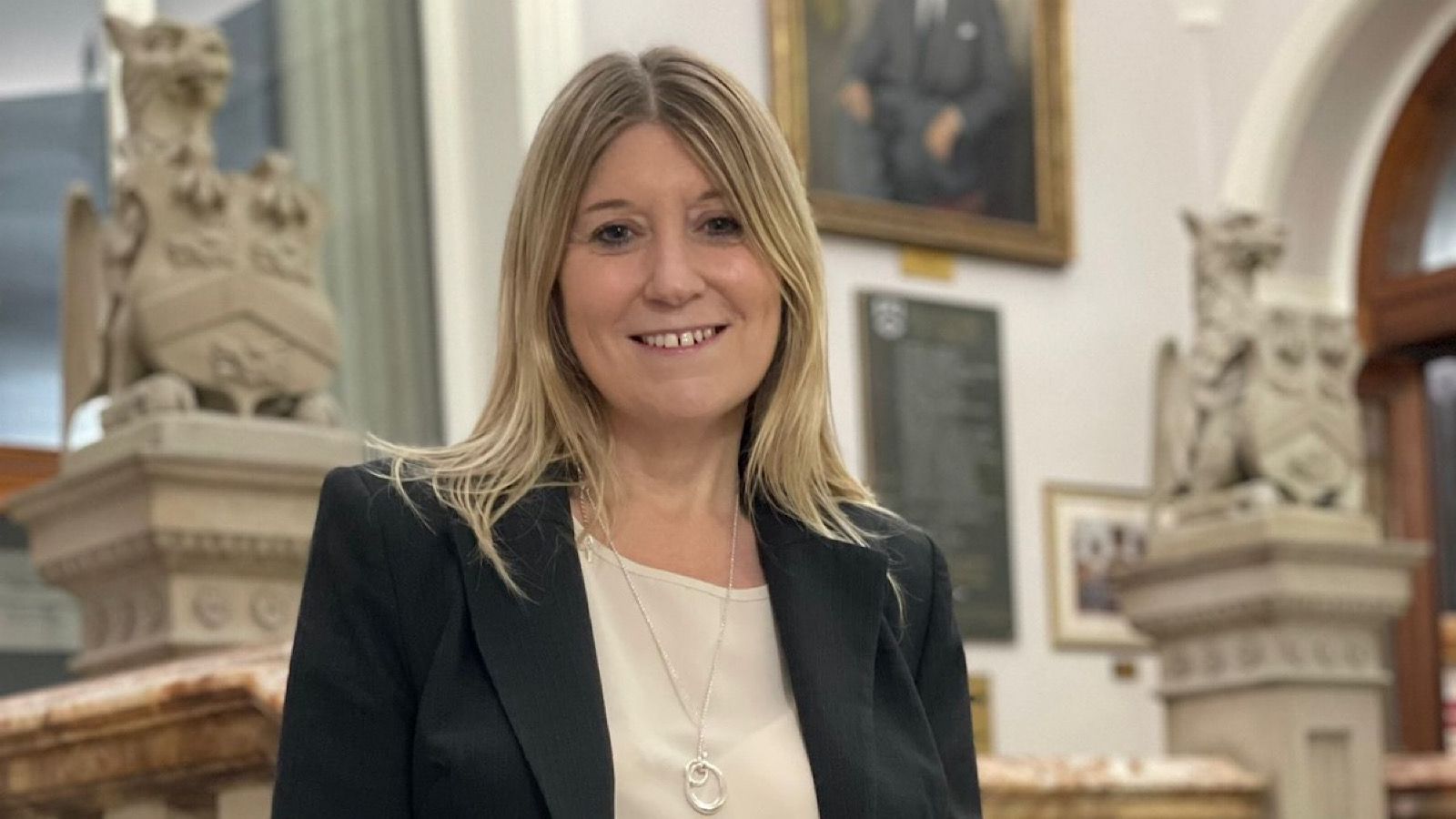 A woman with long blonde hair is smiling, dressed in a black blazer and white blouse, standing indoors in front of ornate stone sculptures.