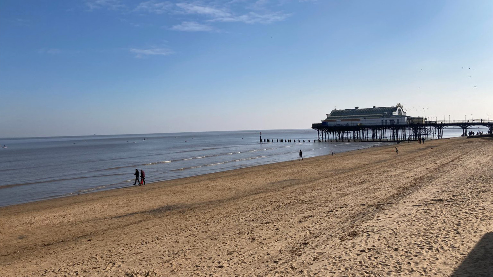 A sandy beach with a pier extending over calm waters. Two people walk along the shoreline under a clear blue sky, as Grimsby Town Foundation and GTFC join the Great British Beach Clean nearby.