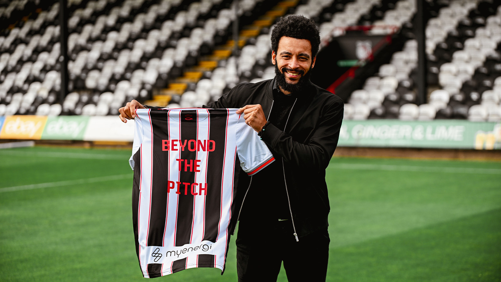 Man holding a striped soccer jersey with "Beyond the Pitch" written on it, standing on a football field.