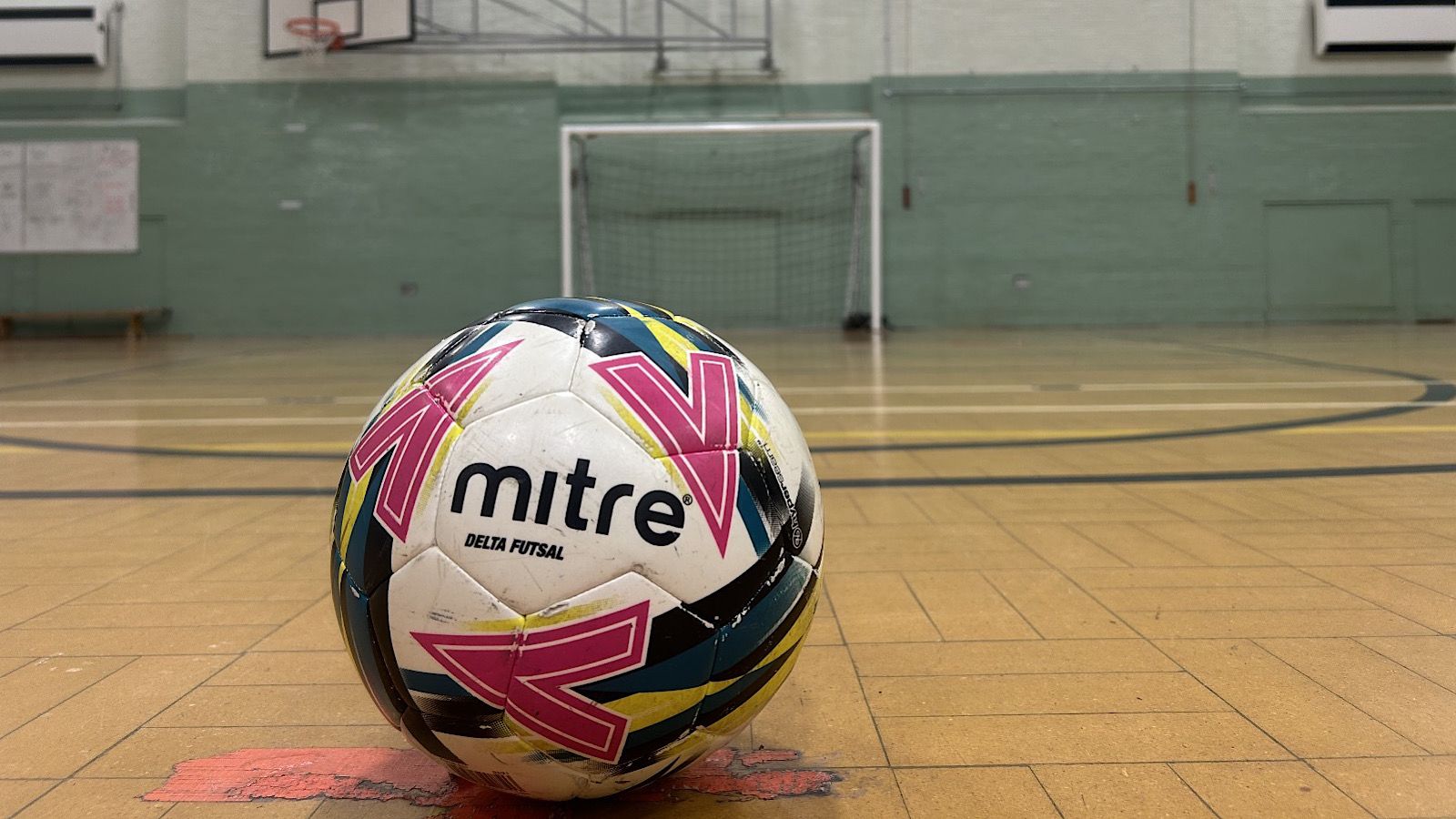 A futsal ball with pink and black patterns is on a gym floor, positioned centrally with the goal visible in the background.