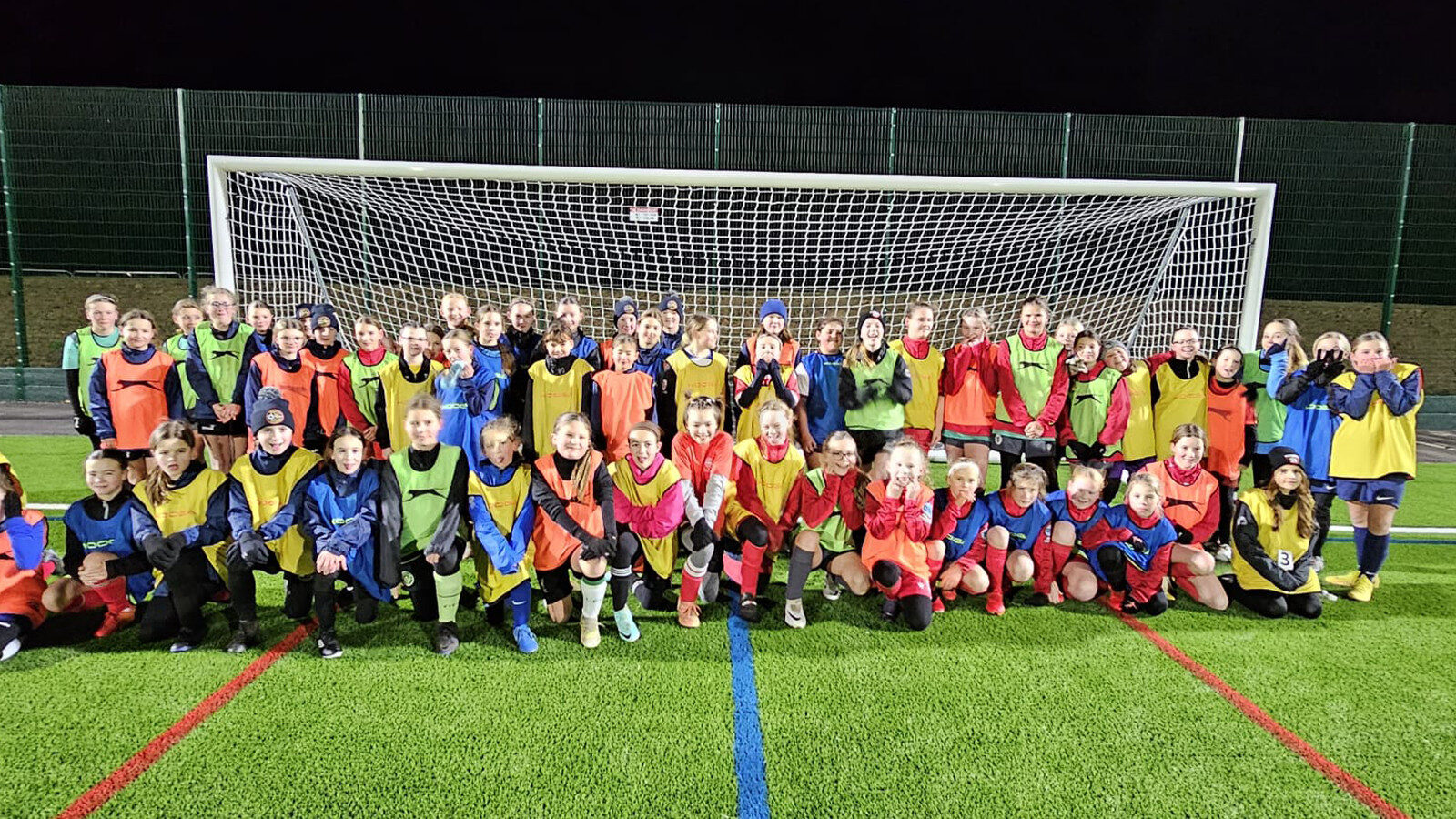 A large group of children in colorful sports bibs pose together in front of a soccer goal on an artificial turf field at night.