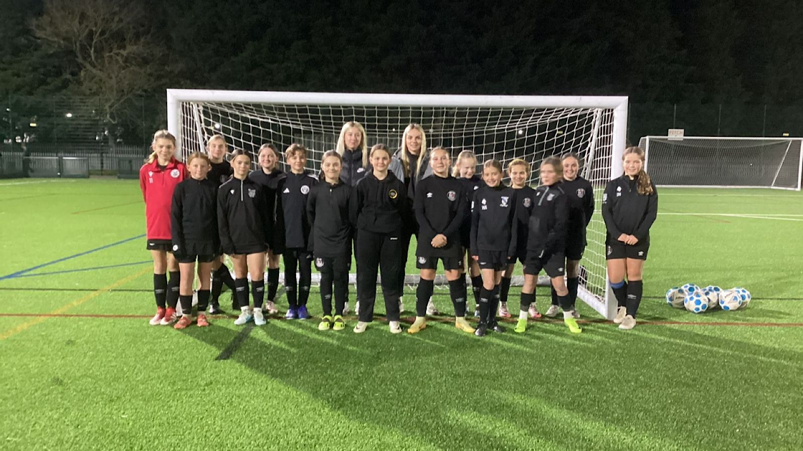 A group of young girls in black sports uniforms stand in front of a soccer goal on a field at night, next to a pile of soccer balls.