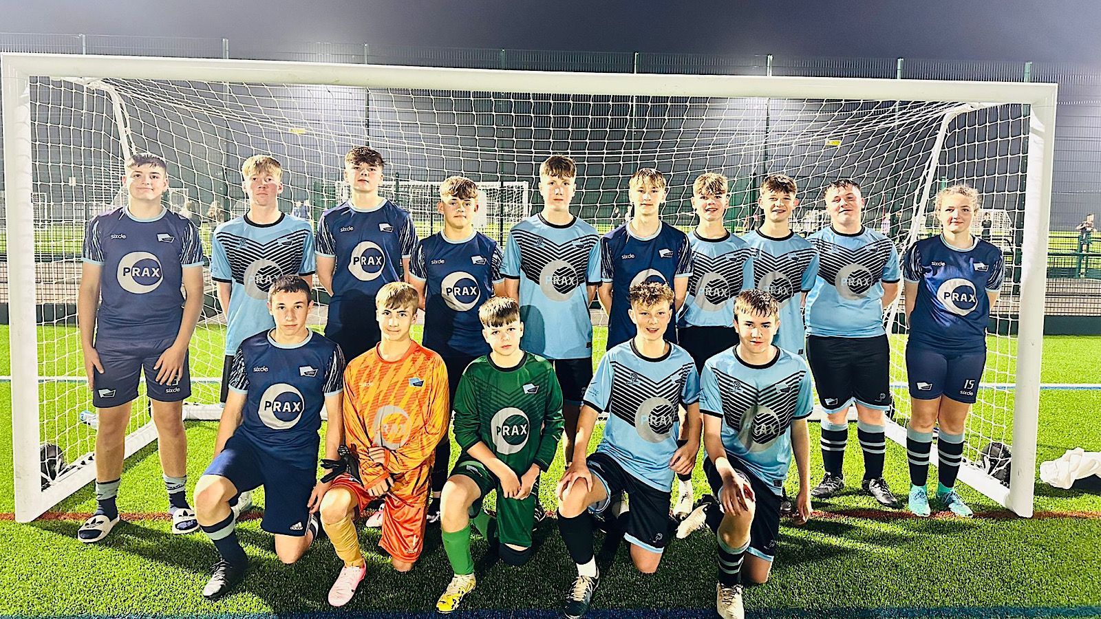 A soccer team poses in front of a goal on a field at night. The players are wearing matching blue and black uniforms, with two in goalkeeper kits.