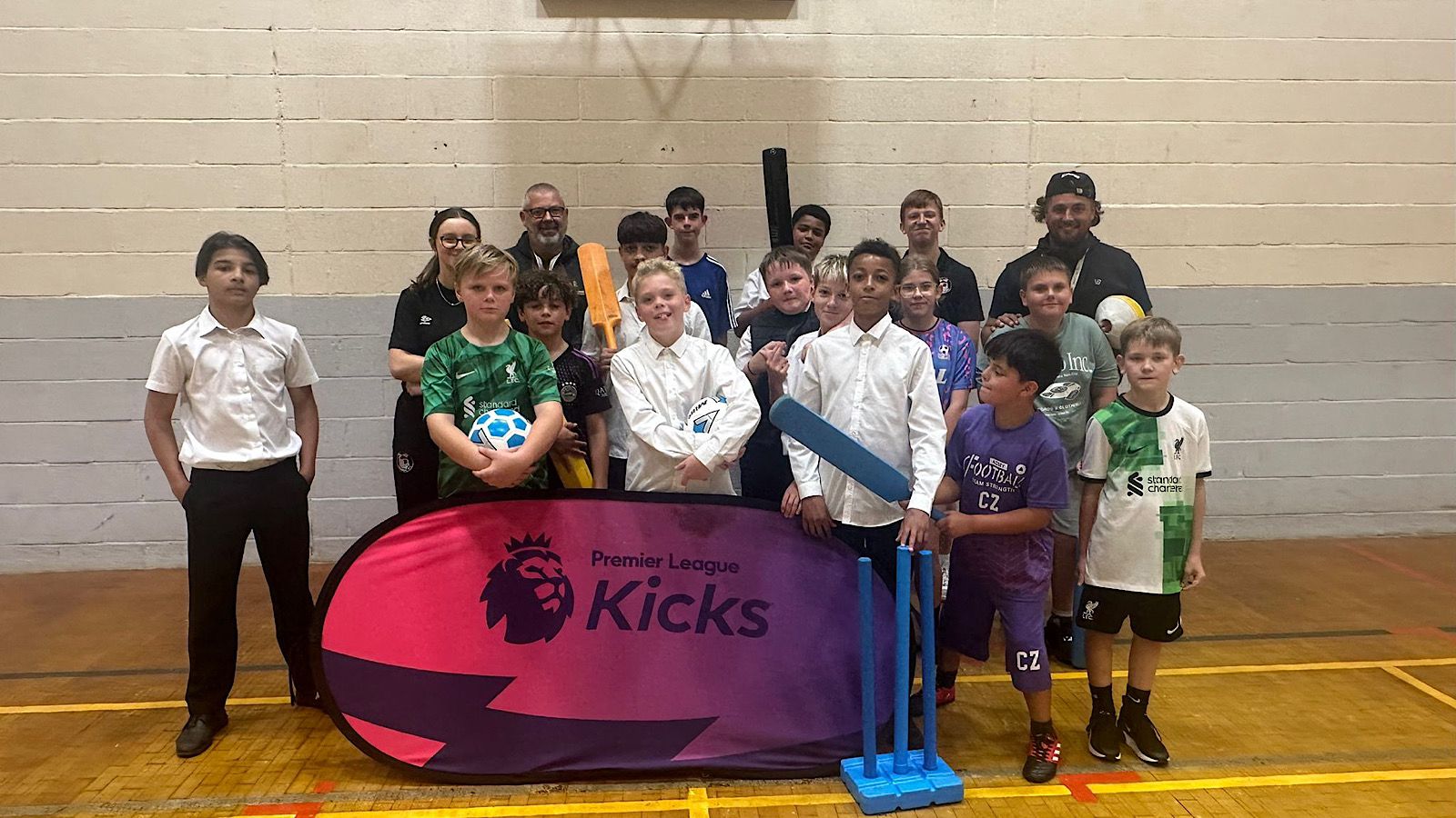 A group of children and adults pose indoors with sports equipment. A "Premier League Kicks" banner is displayed in front.
