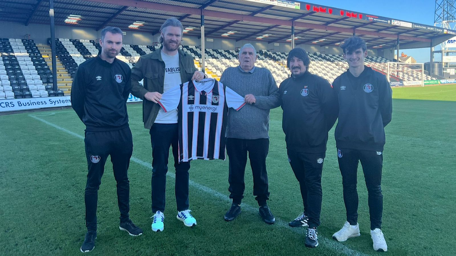 Five people stand on a football field, holding a black and white striped jersey with a "myenergi" logo at the center. The stadium seating is visible in the background.