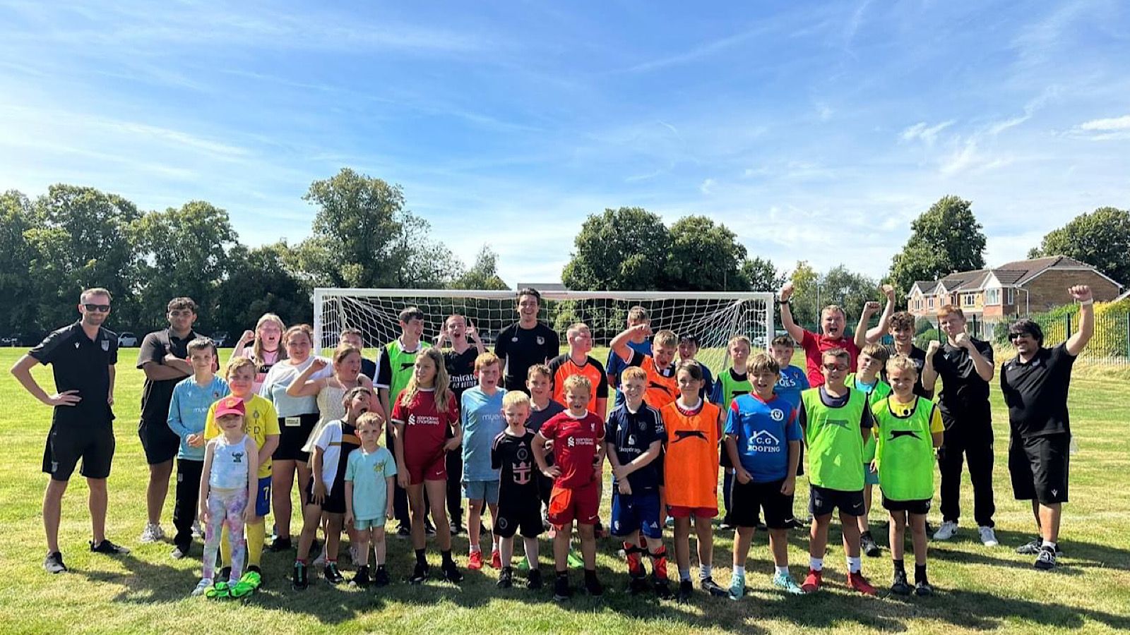 A group of children and adults pose in front of a soccer goal on a grassy field, with some raising their hands. The sky is clear, and trees and houses are visible in the background.