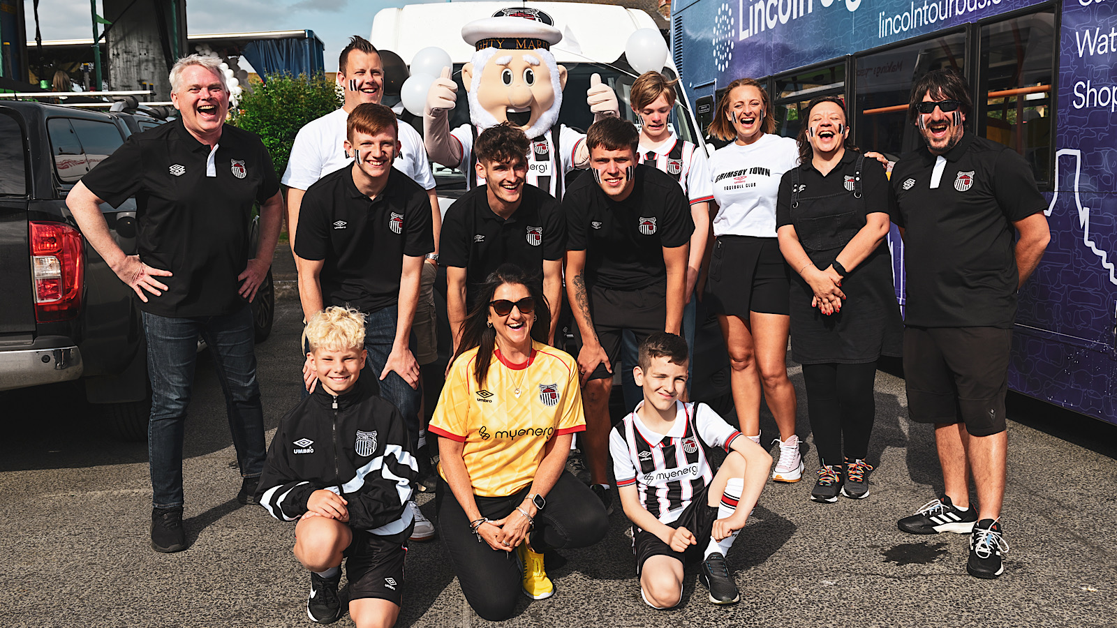 A group of people wearing matching sports attire pose together outdoors with a mascot. Some are standing, others are kneeling, and all are smiling towards the camera.