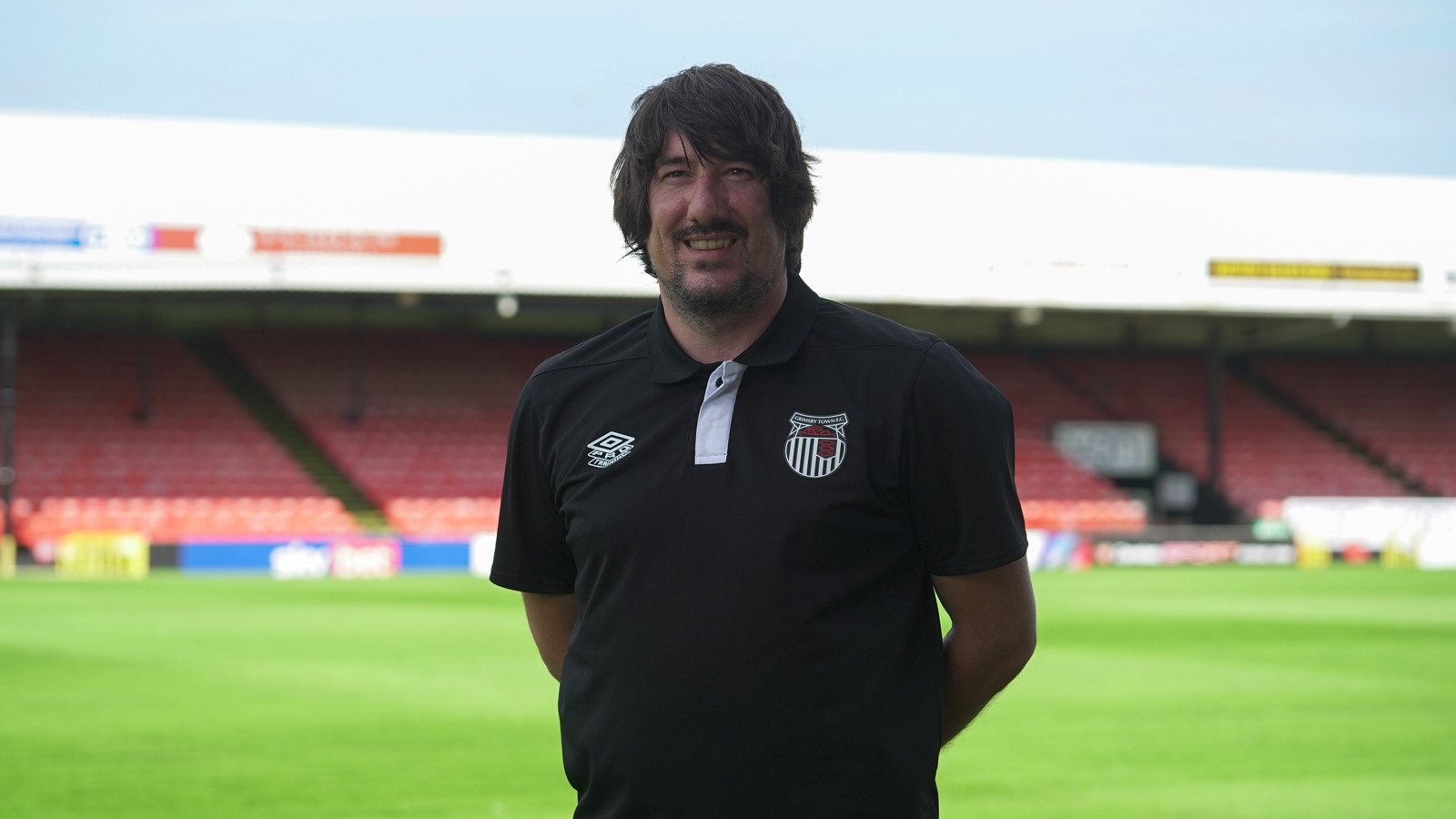 A man with shoulder-length dark hair and a beard, wearing a black polo shirt with a sports team's emblem, stands on a soccer field with empty red and white stands in the background, proudly holding his FA Award for Jason.
