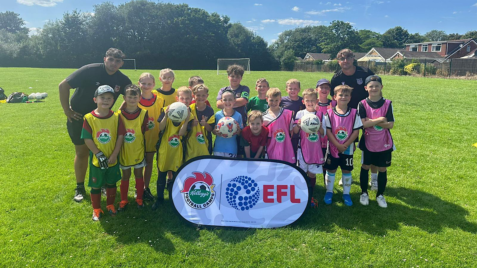 A group of children and two adults stand on a grassy field, holding soccer balls. They are wearing colored sports vests and are gathered behind a banner with the logos of the EFL and another organization.