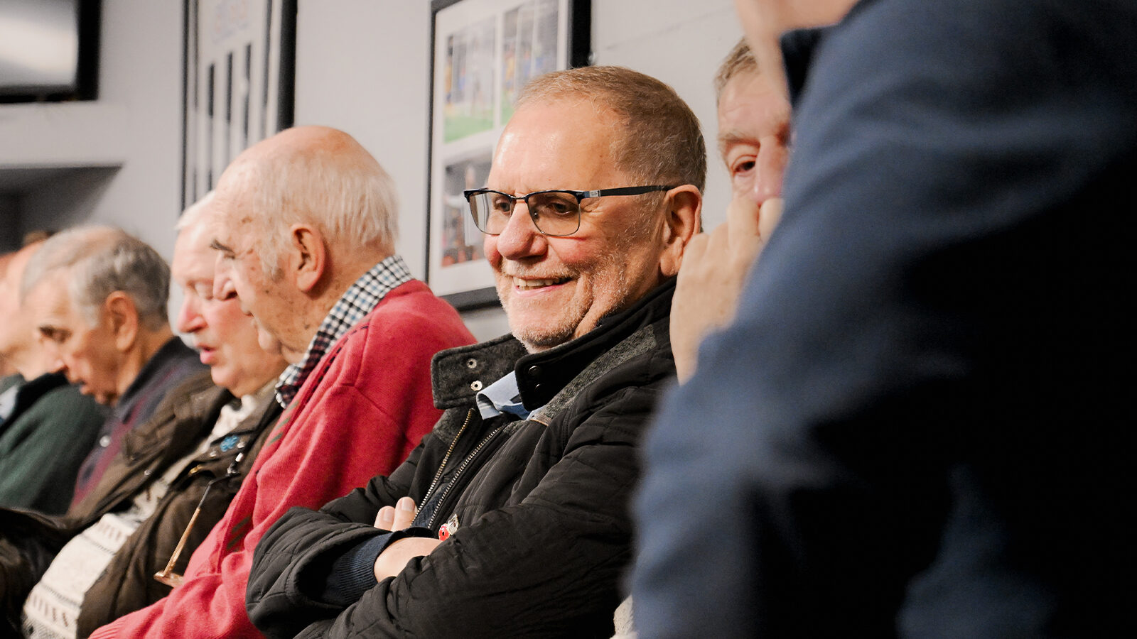 A group of older men sit in a row indoors, wearing casual clothing. One man in the center, wearing glasses, smiles with his arms crossed.