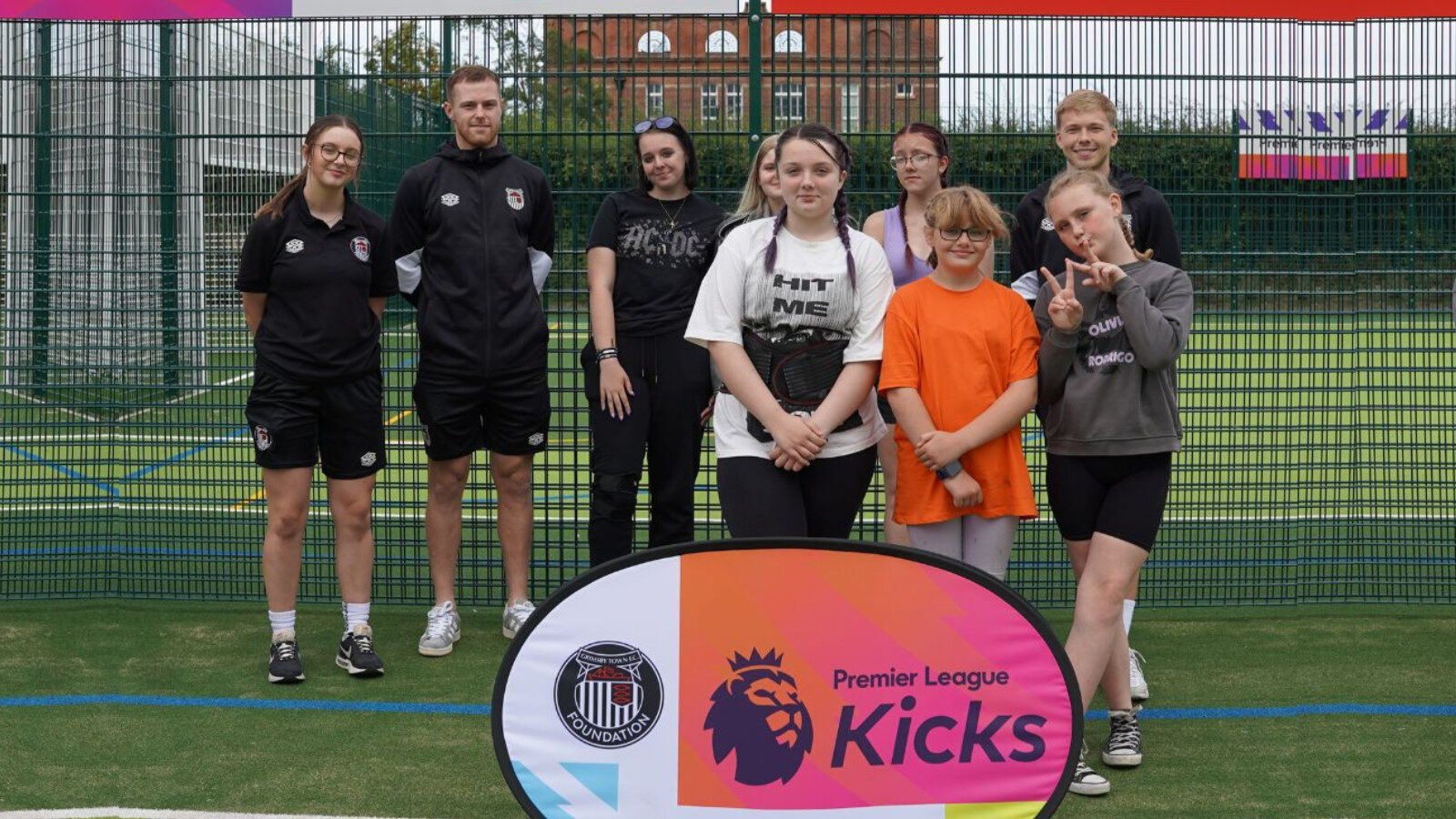 A group of young people and adults pose on an outdoor sports court behind a "Premier League Kicks" banner. Some are wearing sportswear; two girls stand in front, one making a peace sign.
