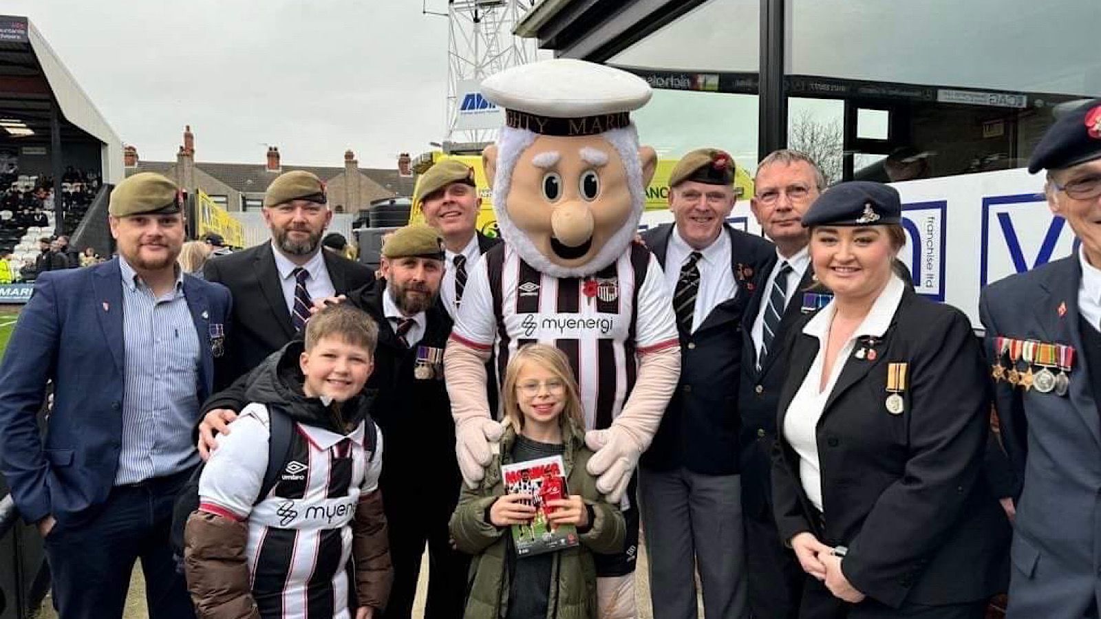 Group of people, including a mascot in a sports uniform, posing for a photo with some members in military-style attire and children holding a magazine.