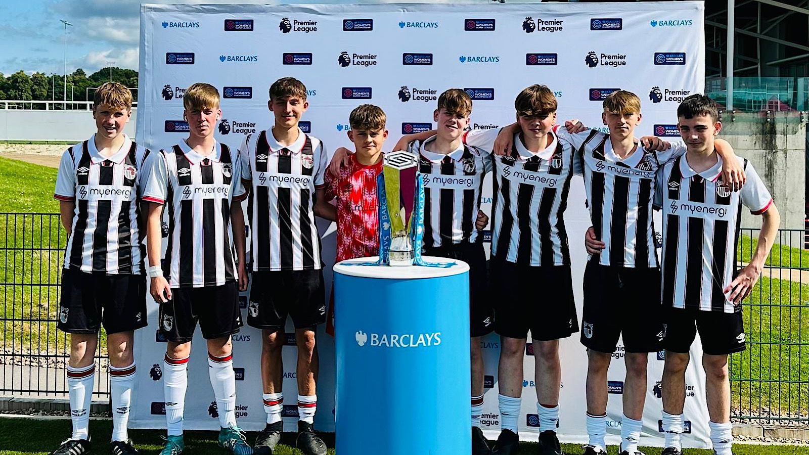 A group of seven boys in black and white striped jerseys stand next to a Barclays trophy on display, posing for a photo in front of a Premier League and Barclays branded backdrop.