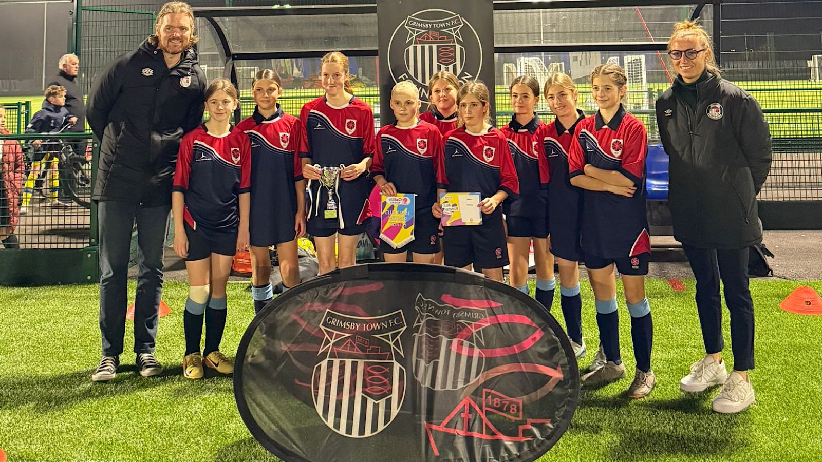 A youth soccer team poses on a field with two coaches. The players hold colorful papers and envelopes. A banner with a logo is displayed in front of them.