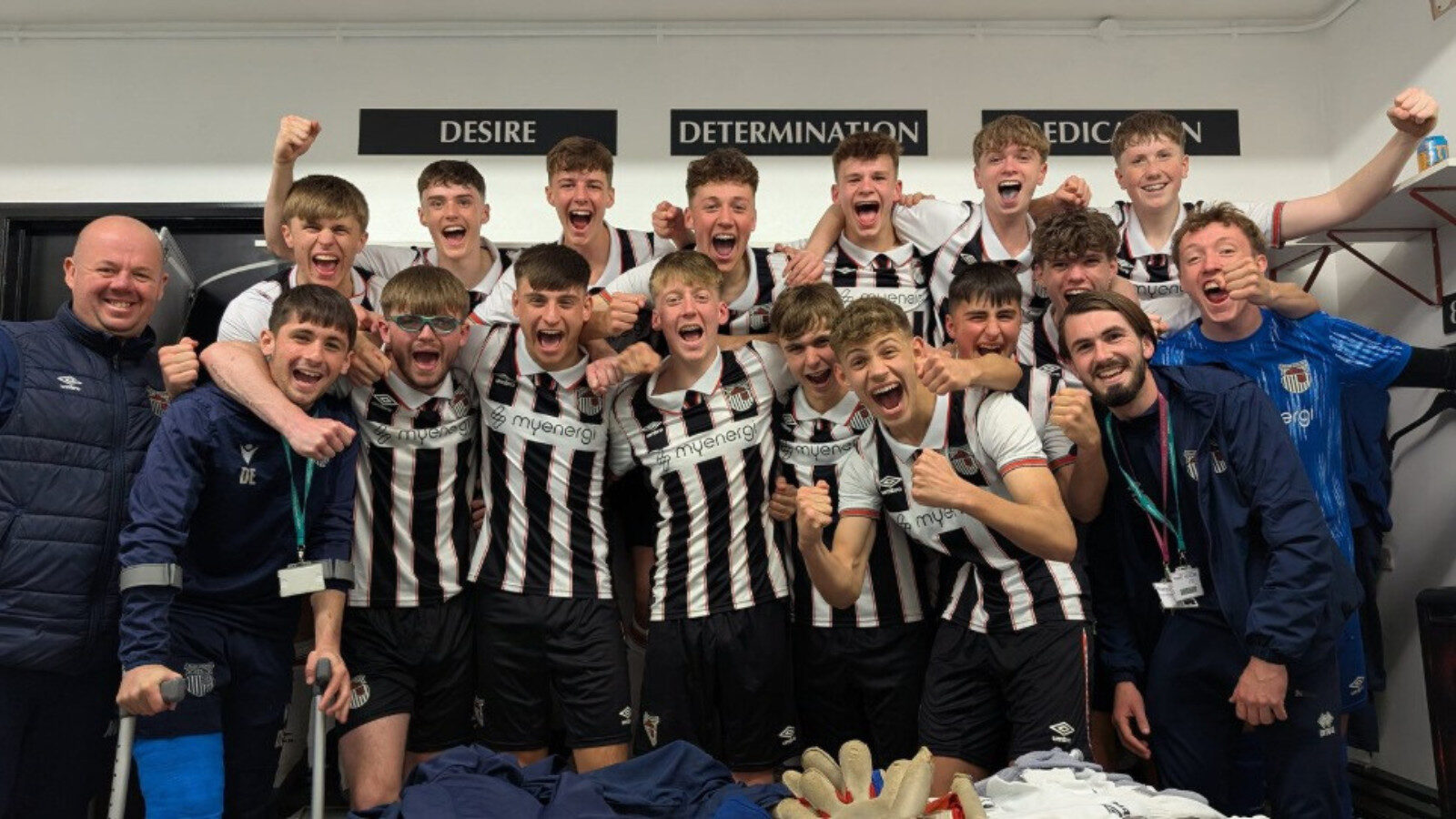 A soccer team in black and white uniforms poses together in a locker room, celebrating with smiles and raised arms in front of motivational words on the wall.