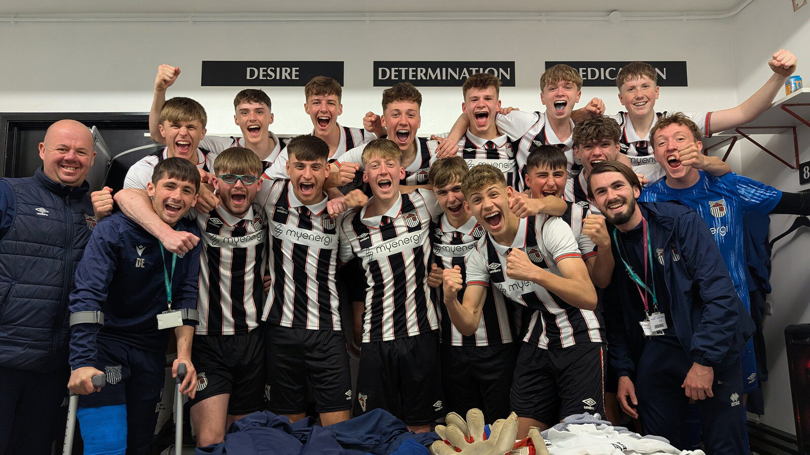 A soccer team poses excitedly in a locker room, wearing black and white uniforms. Three coaches stand with them. Motivational words are visible on the wall behind.