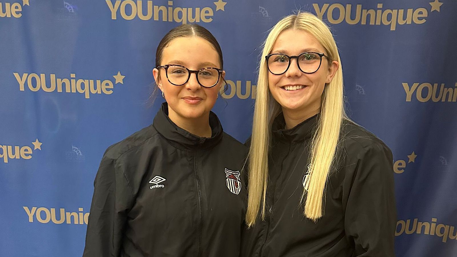 Two people in black jackets and glasses stand in front of a blue backdrop with "YOUnique" written on it.