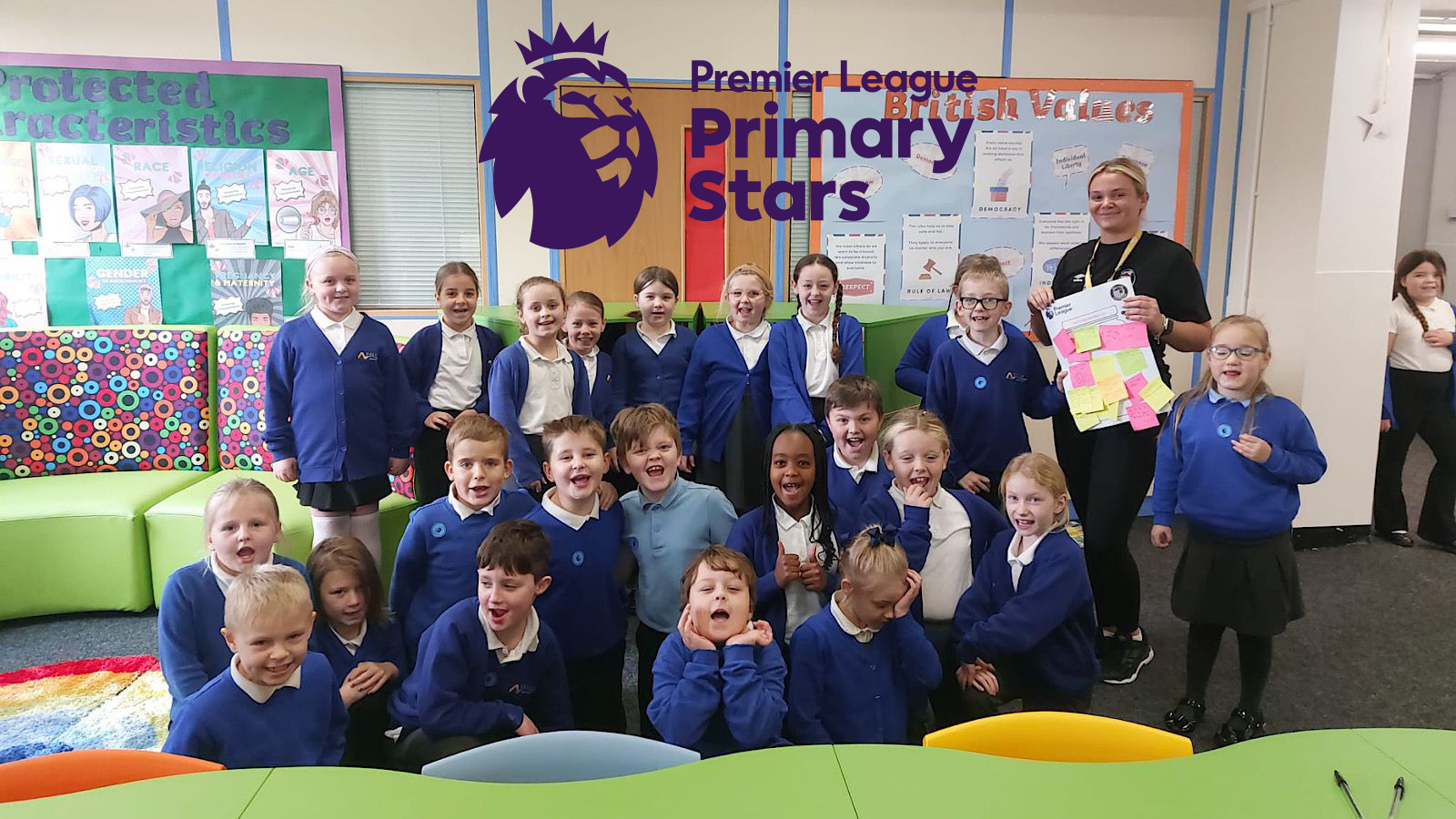 A group of schoolchildren and a teacher pose in a classroom. They're under a Premier League Primary Stars logo and holding colorful paper notes.