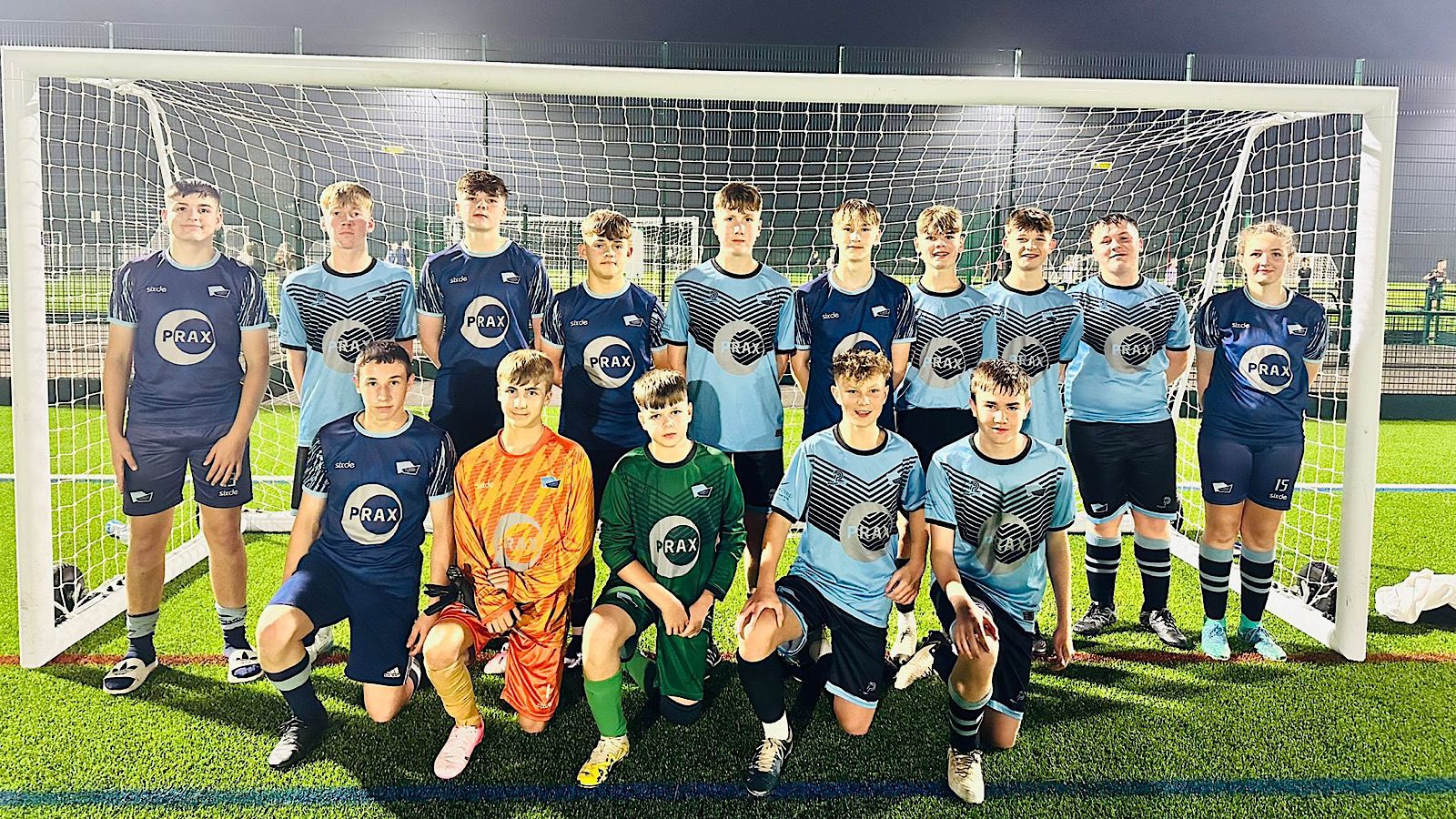 A youth soccer team poses in front of a goal on a turf field at night. The team consists of 15 players wearing blue and light blue uniforms. Two players in goalie uniforms stand in front.