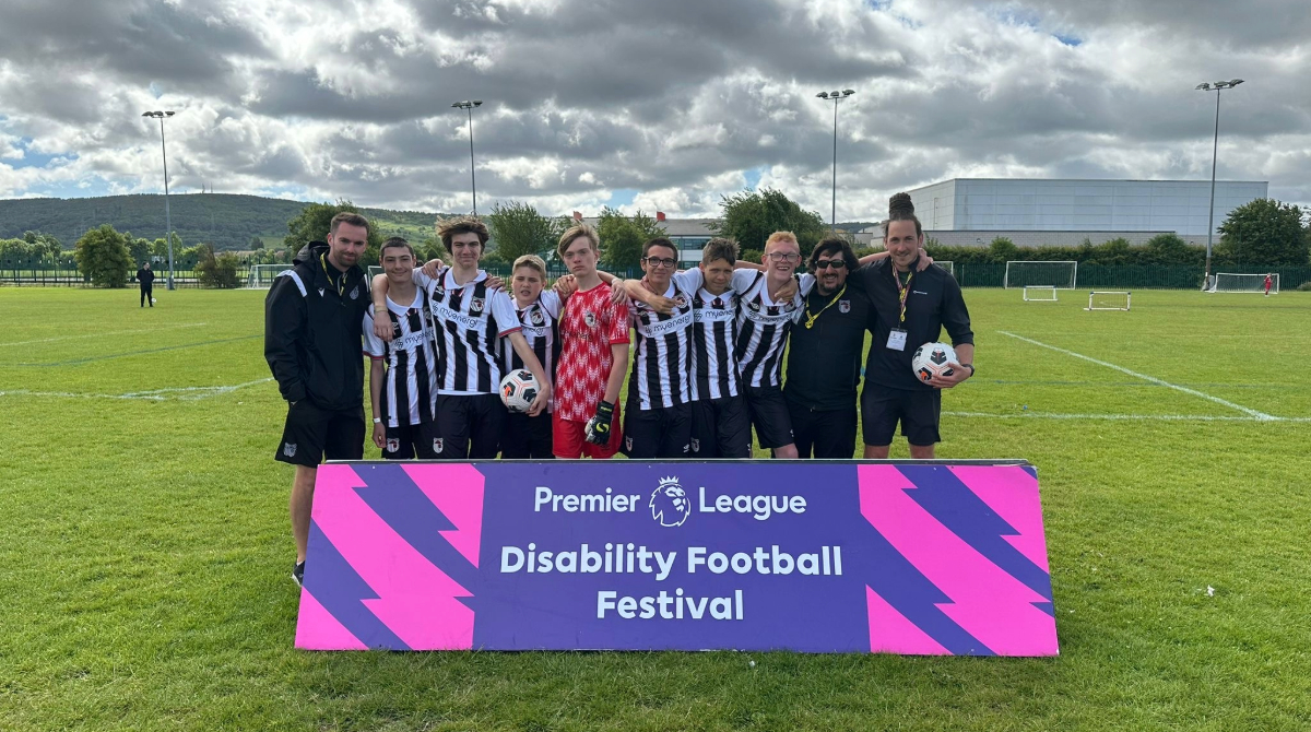 A football team stands on a grass field behind a sign that reads "Premier League Disability Football Festival," with eight players in jerseys and two staff members in black jackets.