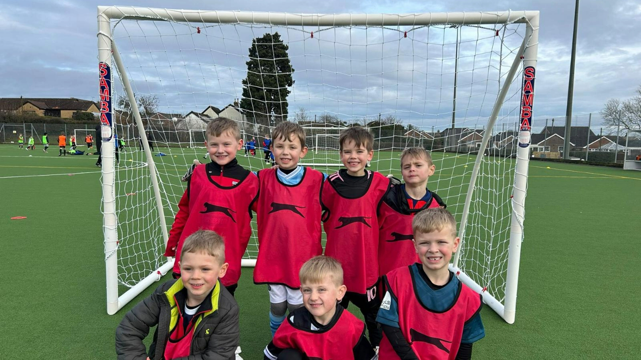 A group of seven boys, wearing red training bibs, stand in front of a soccer goal on an outdoor field. Other players are visible in the background.