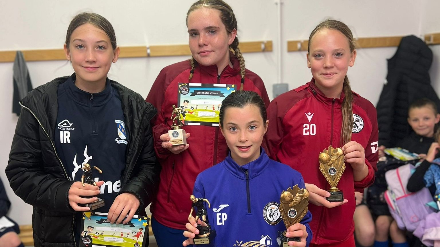 Four young girls dressed in sportswear stand in a locker room holding trophies and certificates.