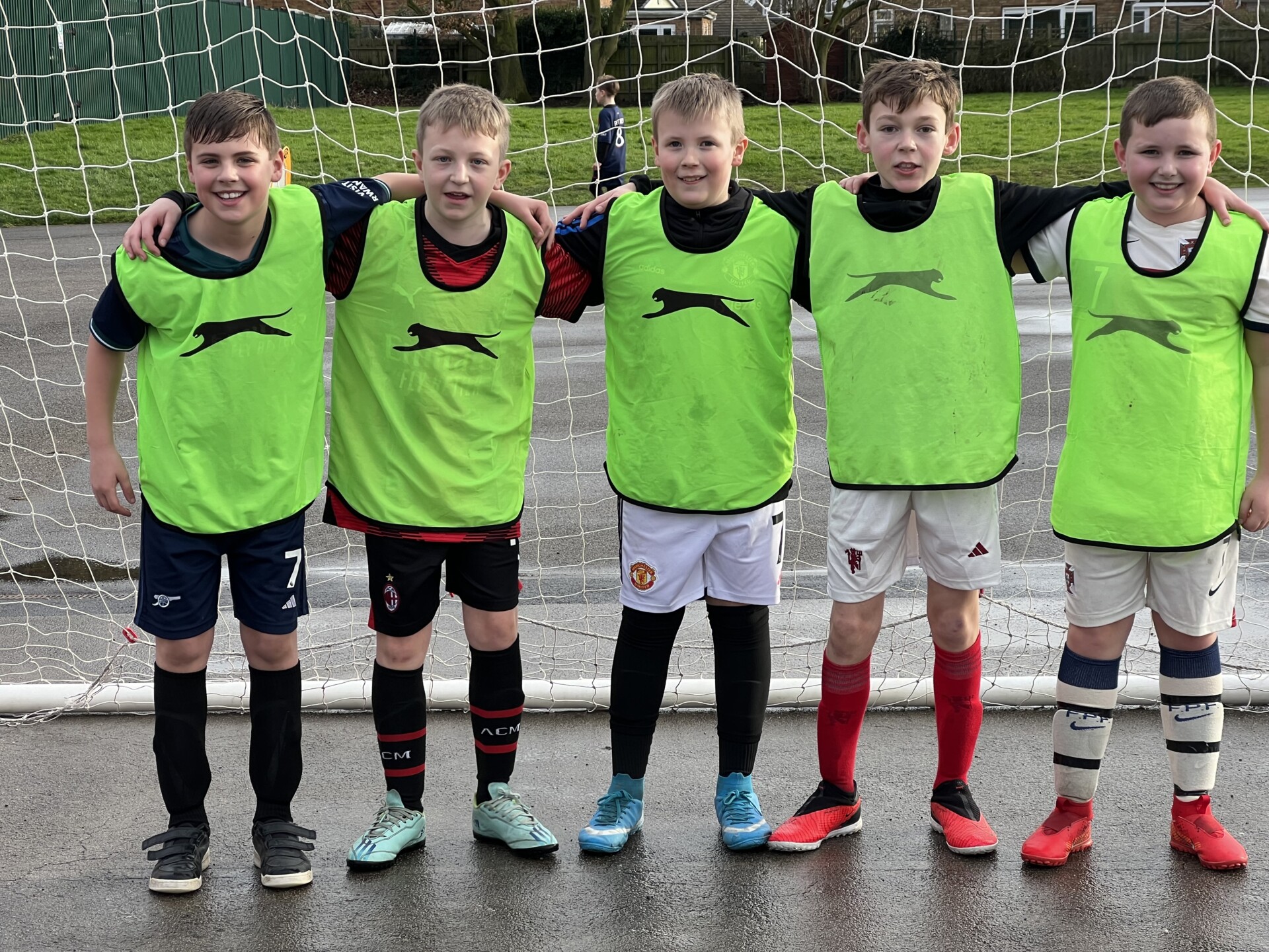 Five young boys in soccer attire and green pinnies stand arm in arm in front of a soccer goal on a paved surface. They are outdoors on a cloudy day.