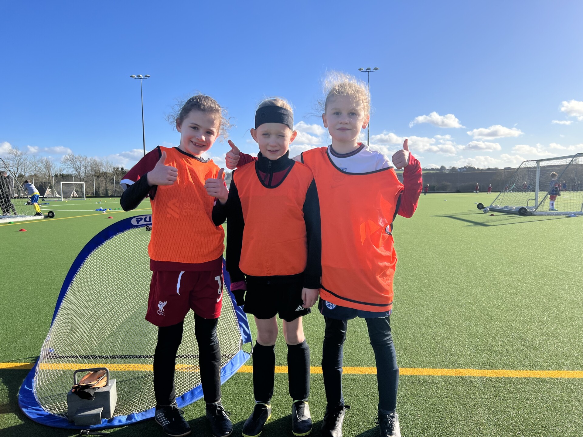 Three children wearing orange training vests stand on an outdoor sports field, smiling and giving thumbs up, with small soccer goals and sports equipment in the background.