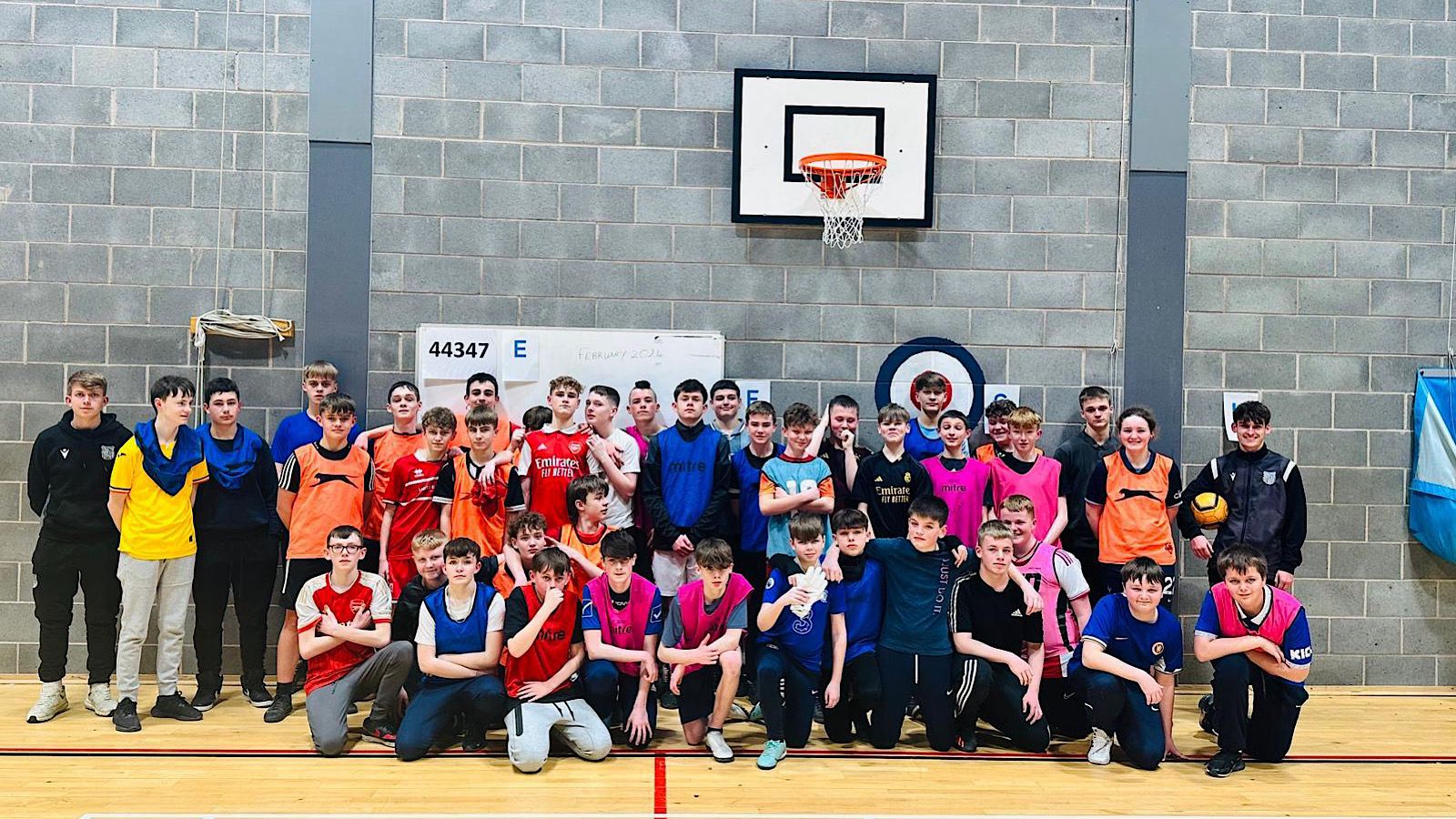 A group of young people pose for a photo in a gymnasium. They are wearing various colored sports bibs, and a basketball hoop is visible in the background.