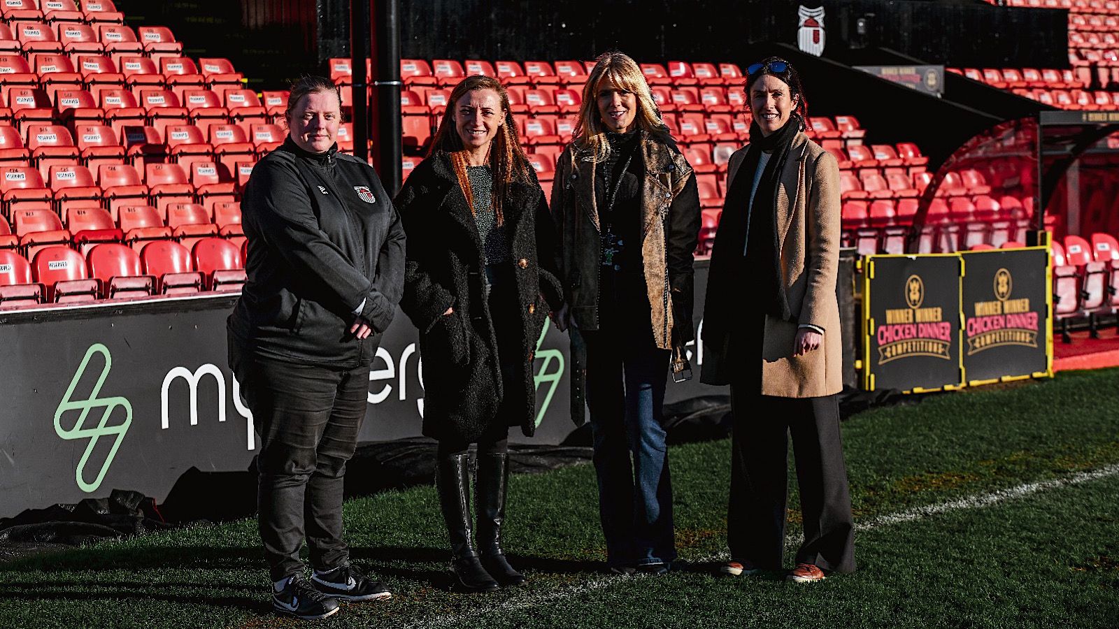 Four people standing on a soccer field in front of red stadium seats, wearing coats and looking at the camera.