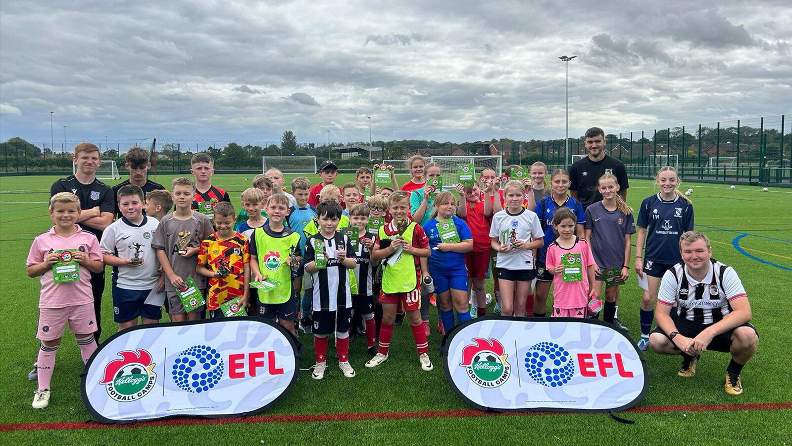 A group of children and a few adults pose on a soccer field, holding certificates, with EFL and club banners in front of them.