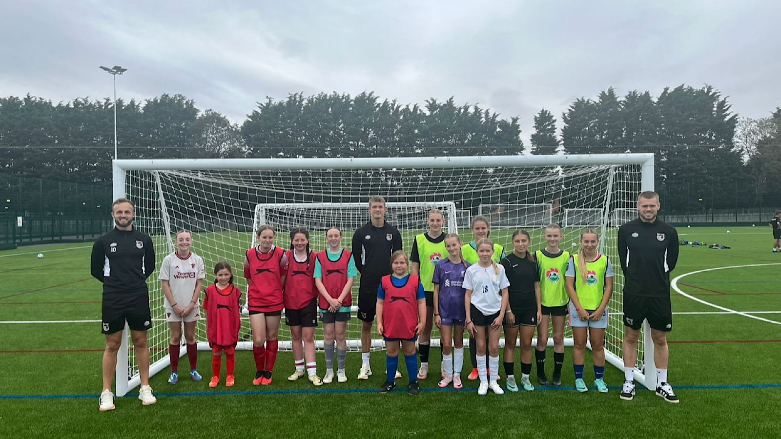A group of soccer players and coaches stand in front of a goal on an outdoor field.