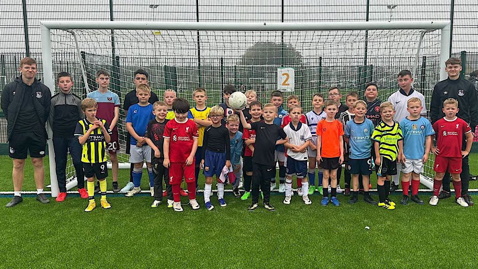 A group of children and adults stand on a soccer field in front of a goal, with some wearing soccer uniforms and one holding a ball.