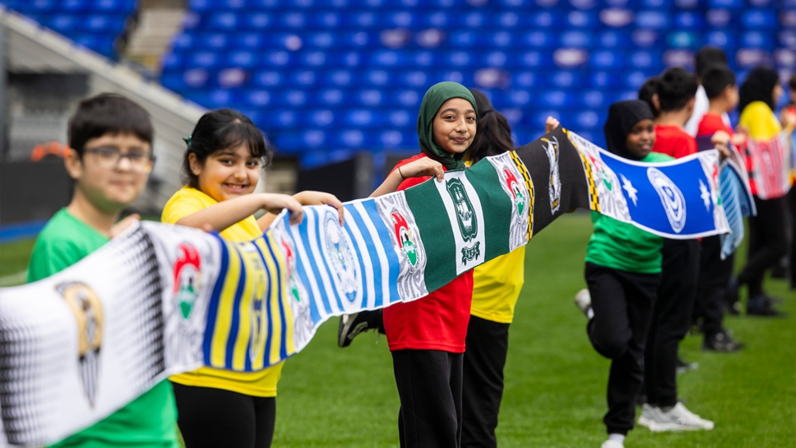 Children holding a row of colorful sports team scarves at a stadium, united by the foundation to deliver Kellogg's football camps.