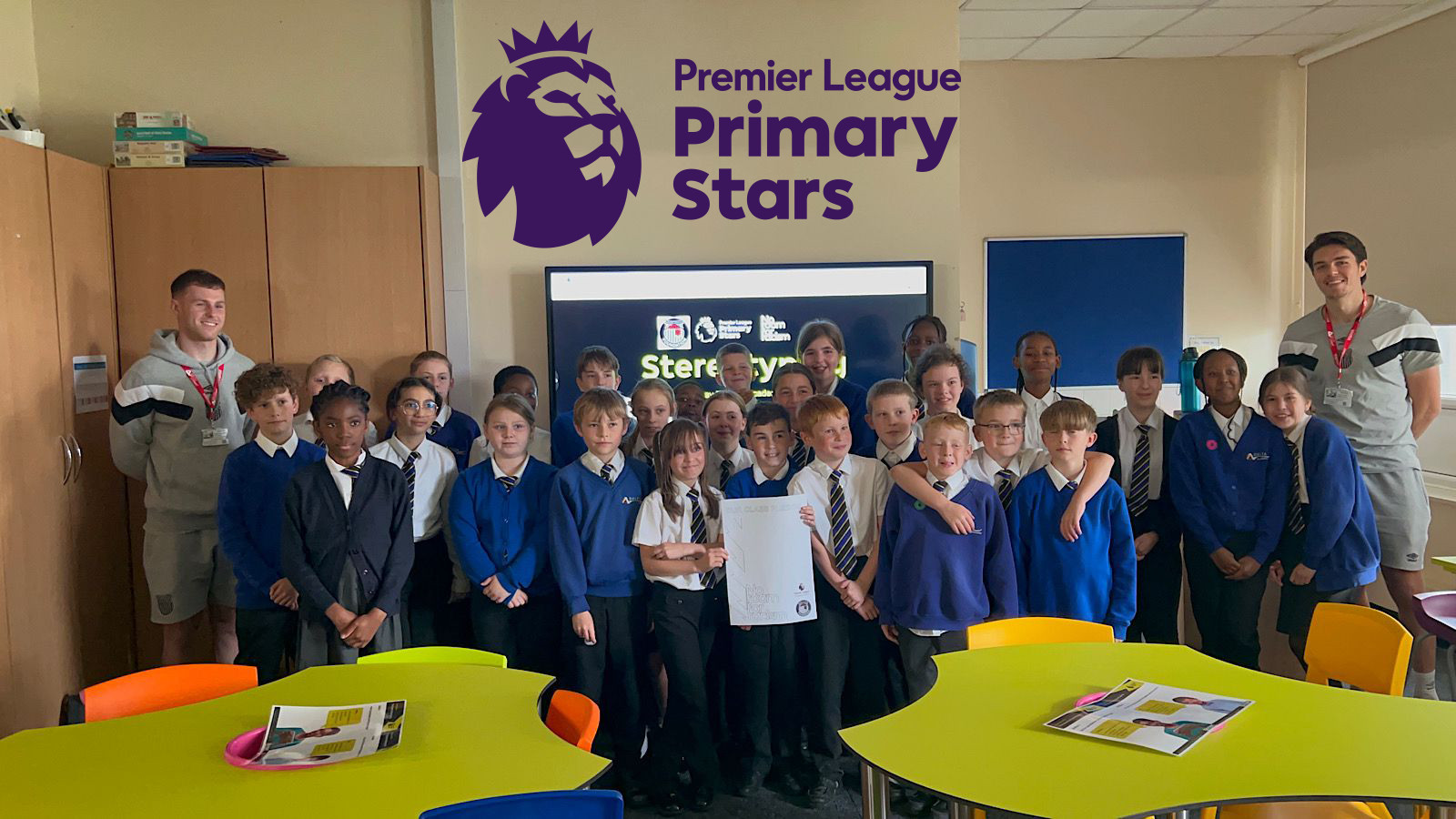 A group of schoolchildren, some in uniforms, and three adults pose in a classroom under a "Premier League Primary Stars" banner.