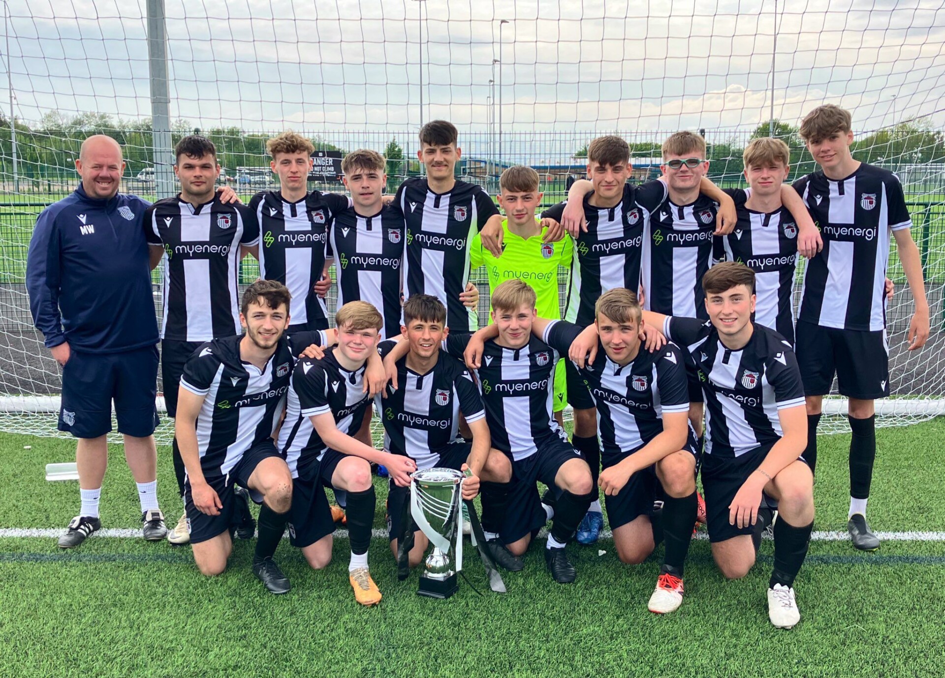 A youth soccer team poses together on a field in black and white uniforms, with a coach standing to the left. They are holding a large trophy in front of a goal net.