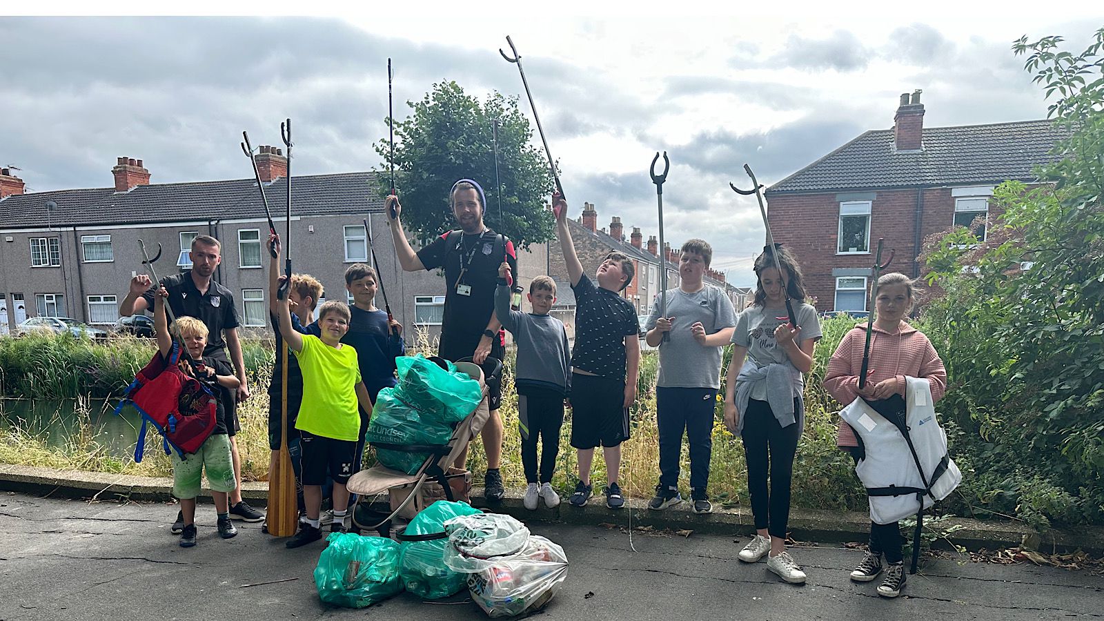 A group of people, including children and adults, stand on a roadside with trash bags and litter pickers, participating in a community cleanup. Houses and trees are in the background.