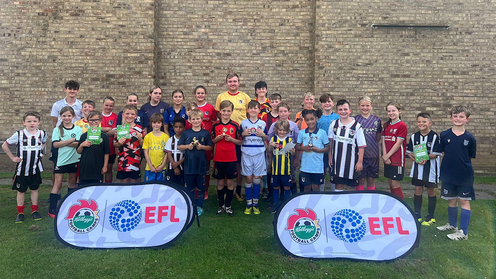 A group of kids in various football uniforms pose for a photo on a grassy field with two EFL banners in front. They are smiling and some are holding trophies. A brick wall is in the background.