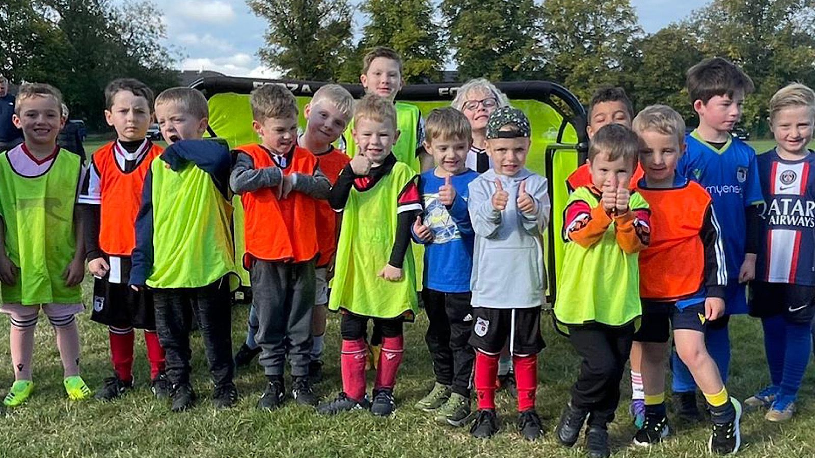 A group of children in sports attire stand on grass, with several wearing orange and green vests, posing for a photo. Trees and a portable sports net are visible in the background.