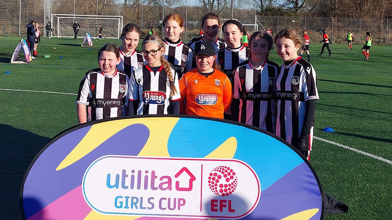 A soccer team of young girls wearing black and white uniforms poses behind a banner reading "Utilita Girls Cup EFL" on a field.