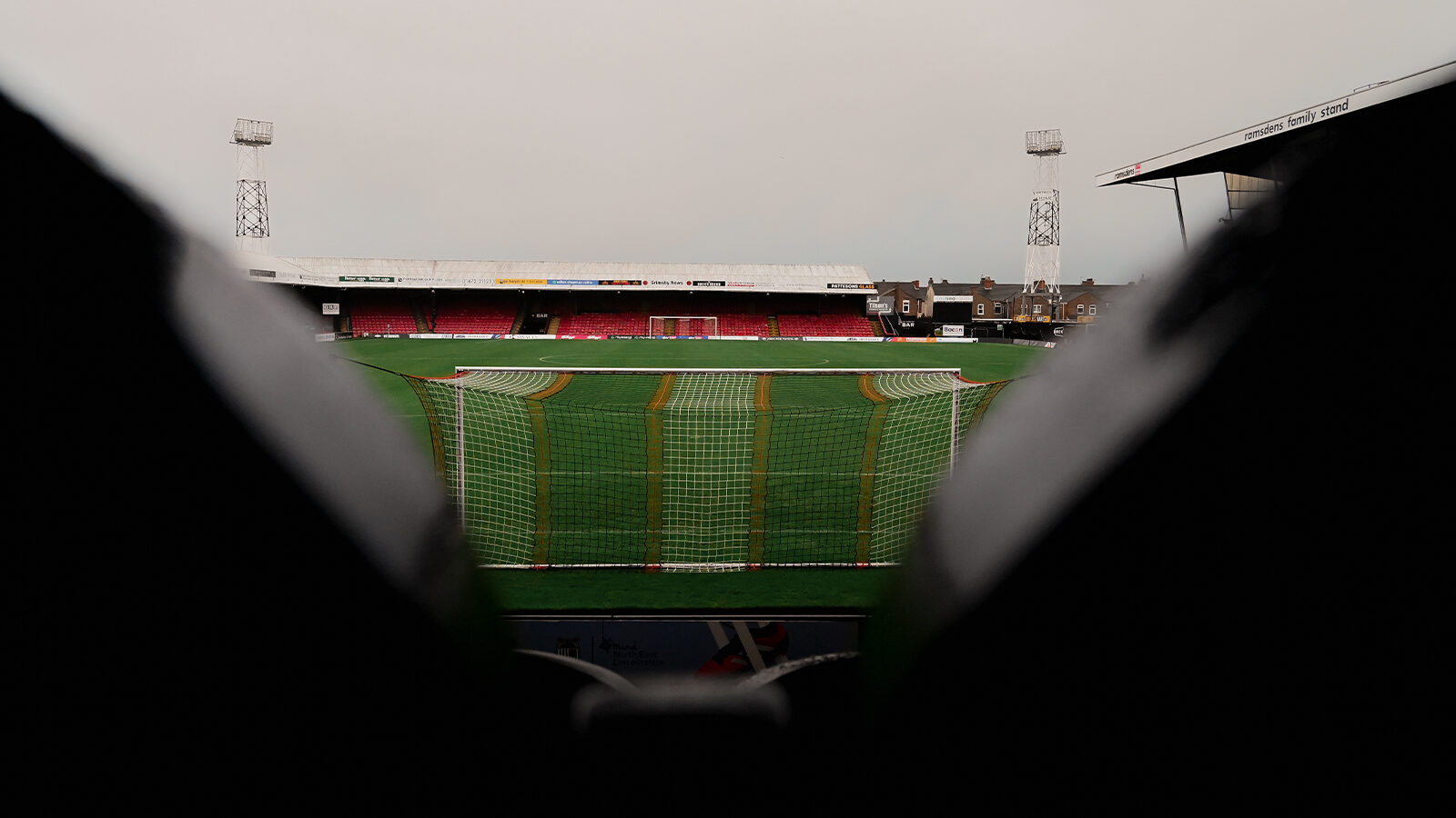 A view of an empty soccer stadium from behind two seats, showing the goalpost, green field, and red stands under an overcast sky.