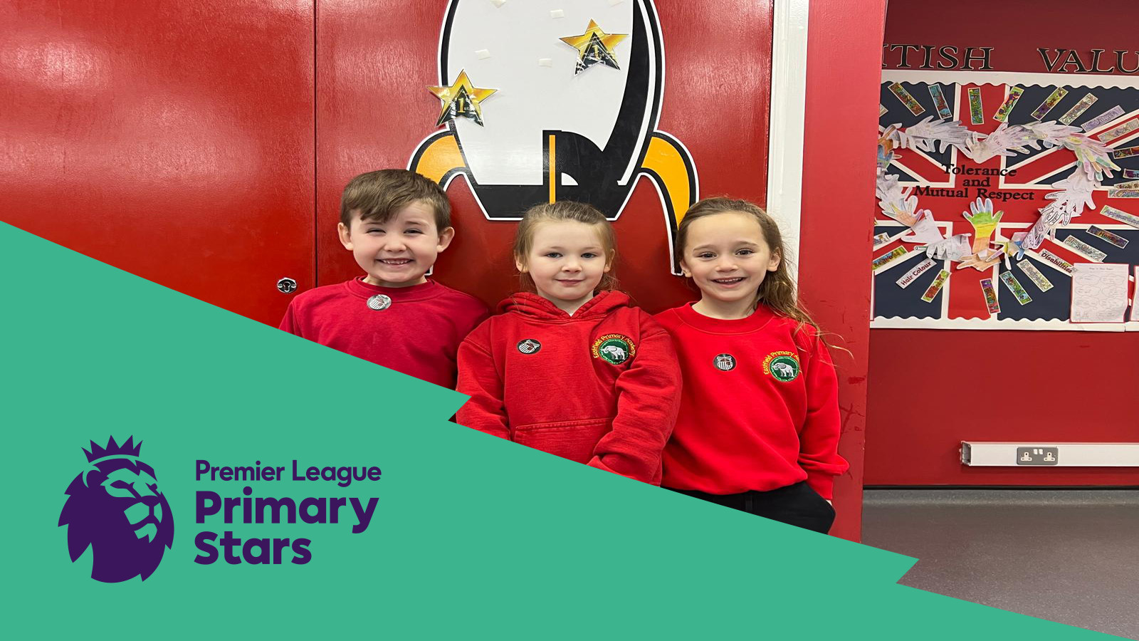 Three children wearing red hoodies stand side by side, smiling in front of a red wall with a penguin decoration. Premier League Primary Stars logo is in the bottom left corner.