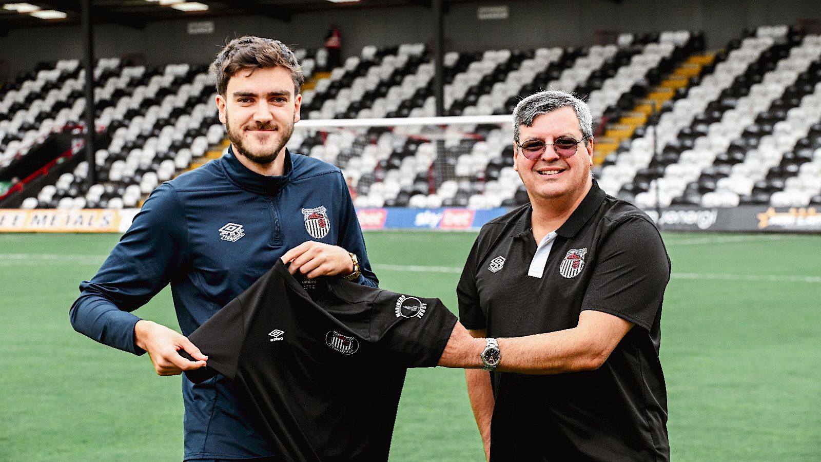 Two people at a sports stadium holding a black sports shirt with a logo.