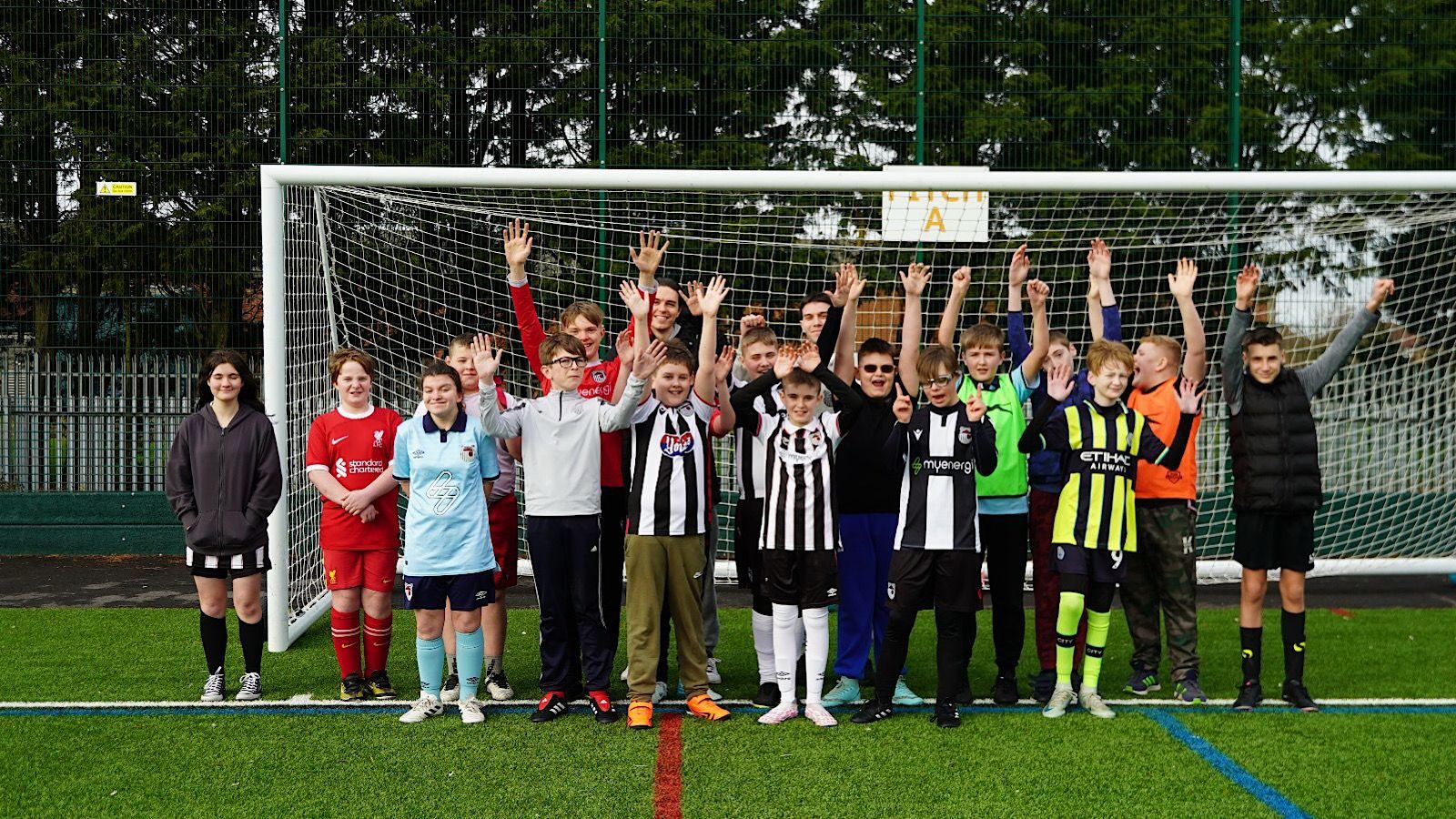 A group of young soccer players in various team uniforms stand on a field in front of a goal, with hands raised.